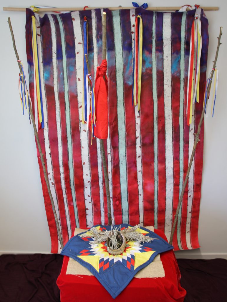 Prayers for My Sisters, "This series is about recognizing that within the issue of MMIWG; the women are cared for spiritually. They are prayed for and ceremonies held for them. The red background pays homage to the work done like "The Red Dress" by Jaime Black which has raised awareness since 2011. The 12 sticks represent the lodges and the ribbons represent the prayers. I have supplied an image of a painting and also the Installation which allows people to interact with the piece." Dawn Marie Marchand.